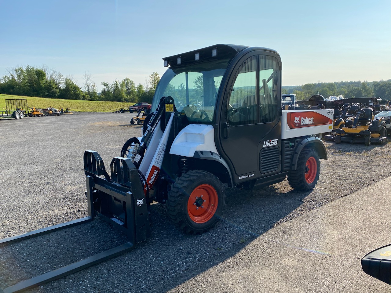 A Bobcat UW56 utility work machine with a forklift attachment is parked on a gravel lot at Greene County Power Equipment in Greenville, NY. The enclosed cab vehicle has a white and black body with red wheels, surrounded by other outdoor power equipment under a clear blue sky.