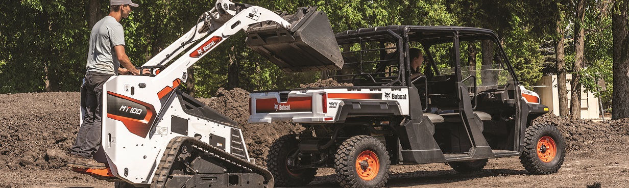 A Bobcat MT100 mini track loader operated by a worker lifts a bucket of soil to load into a Bobcat UV34XL utility vehicle parked nearby. The scene takes place on a dirt worksite surrounded by green trees, with another worker sitting in the utility vehicle.