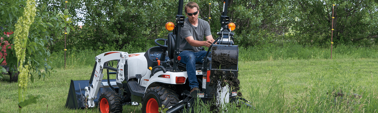 A man operating a Bobcat compact tractor with a backhoe attachment digs into the soil in a grassy field surrounded by trees. The tractor's front loader is also visible, positioned for additional utility in the landscaping or excavation work.