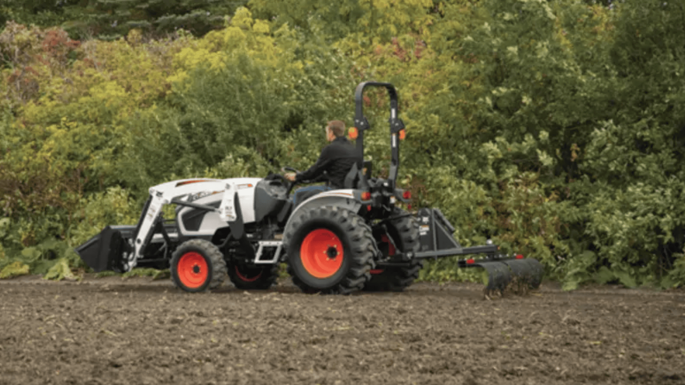 A man drives a 2025 Bobcat® Tine Rake 3PT tractor through a field in Greenville, NY.