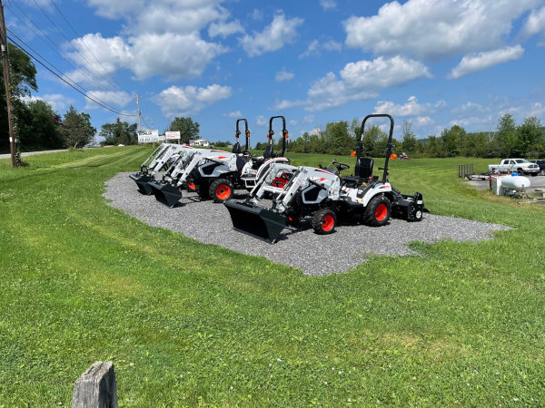 A row of Bobcat compact tractors with front loader attachments is displayed on a gravel lot at Greene County Power Equipment in Greenville, NY. The equipment is set against a backdrop of green grass, trees, and a bright blue sky with scattered clouds.