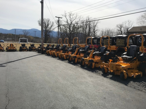 A row of bright yellow Cub Cadet zero-turn mowers is lined up outside Greene County Power Equipment in Greenville, NY. The mowers are parked on a paved lot under a clear sky, with power lines, leafless trees, and distant mountains in the background.