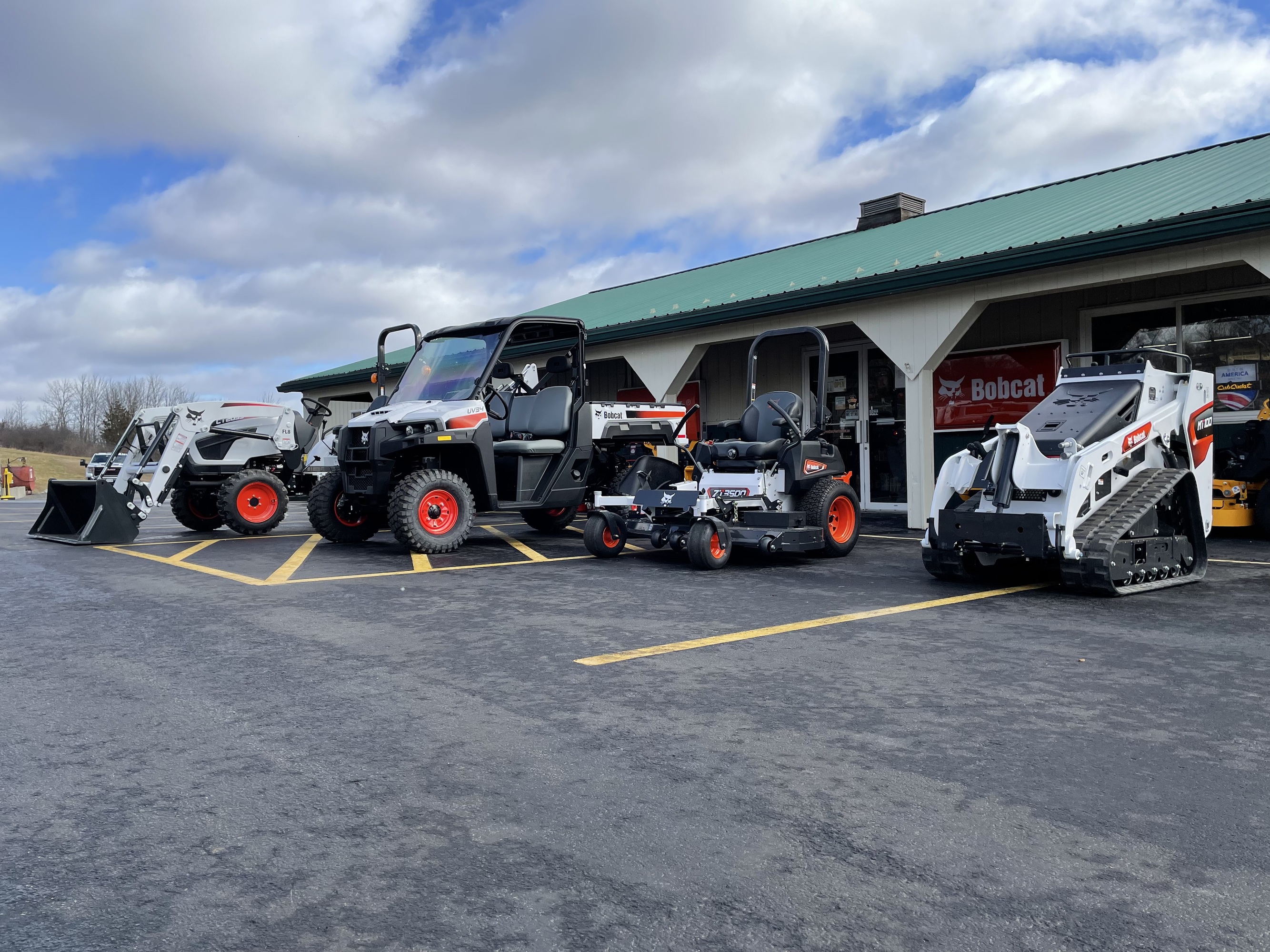 Greene County Power Equipment in Greenville, NY, showcases a lineup of Bobcat outdoor power equipment, including utility vehicles, skid-steer loaders, and zero-turn mowers, parked in front of the store. The building has a green metal roof and white pillars, with a Bobcat sign displayed under a partly cloudy sky.