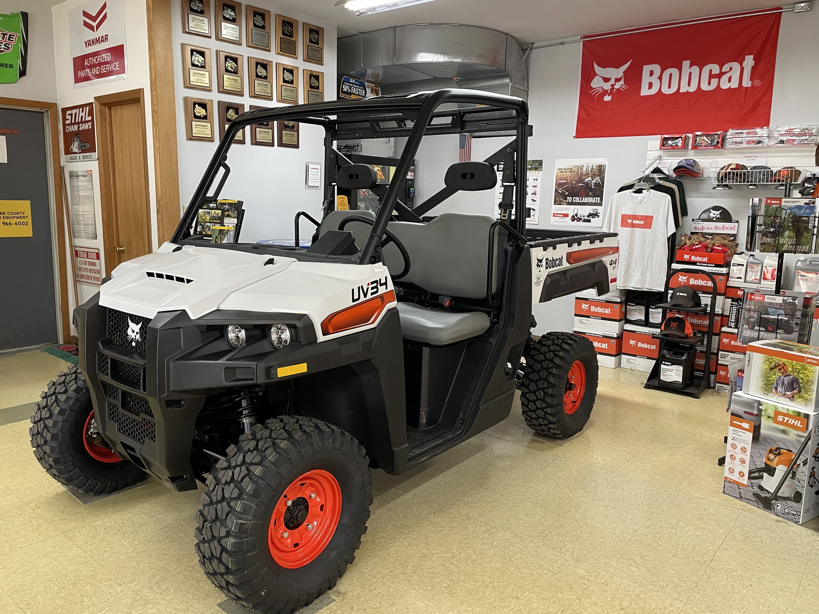 A white Bobcat UV34 utility vehicle with red wheels is displayed inside Greene County Power Equipment in Greenville, NY. The showroom features a Bobcat banner, STIHL products, and various equipment accessories, with award plaques mounted on the wall.